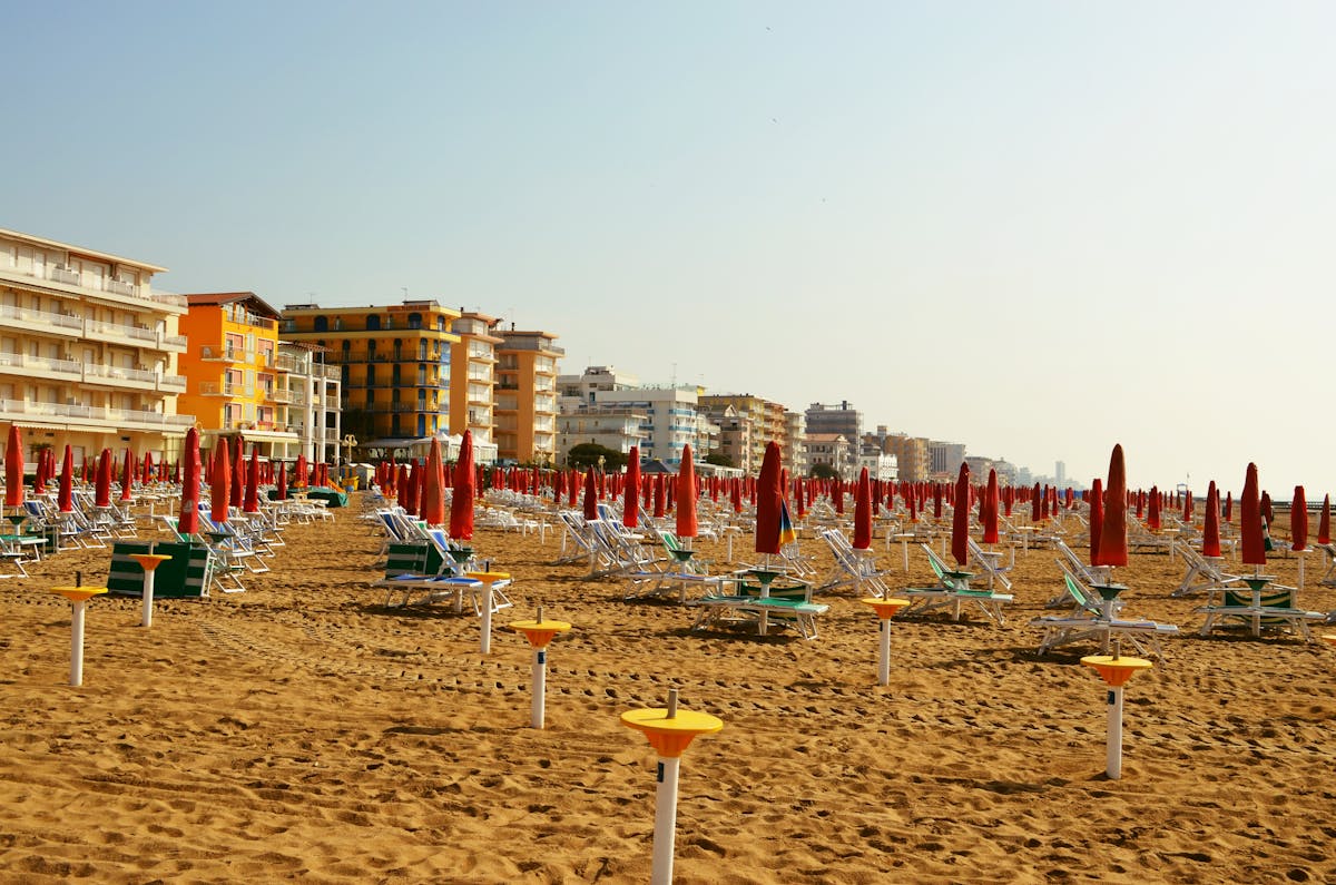 Rows of red and white beach umbrellas on a sandy Italian beach at sunset, with a line of pastel seafront buildings in the background