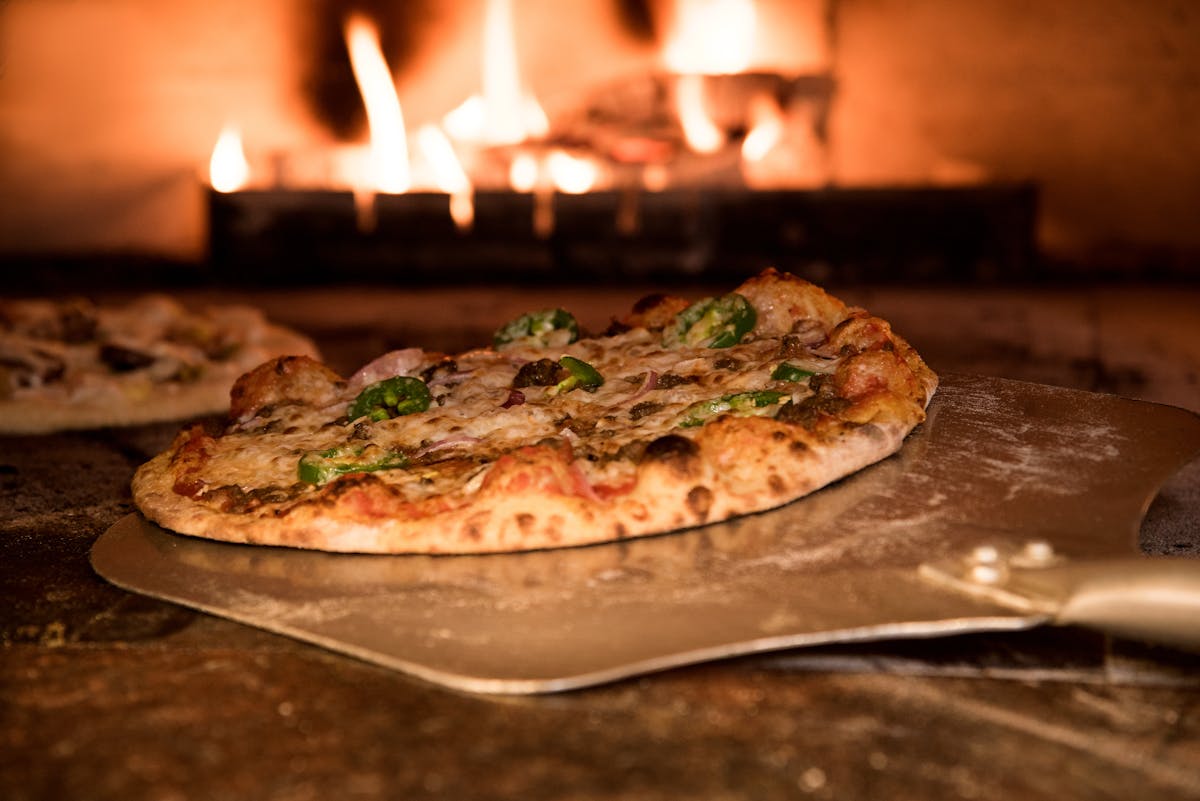 A round wood-fired flatbread on a metal peel in front of a brick oven with live flames, the oven floor and ember-lit chamber visible behind it