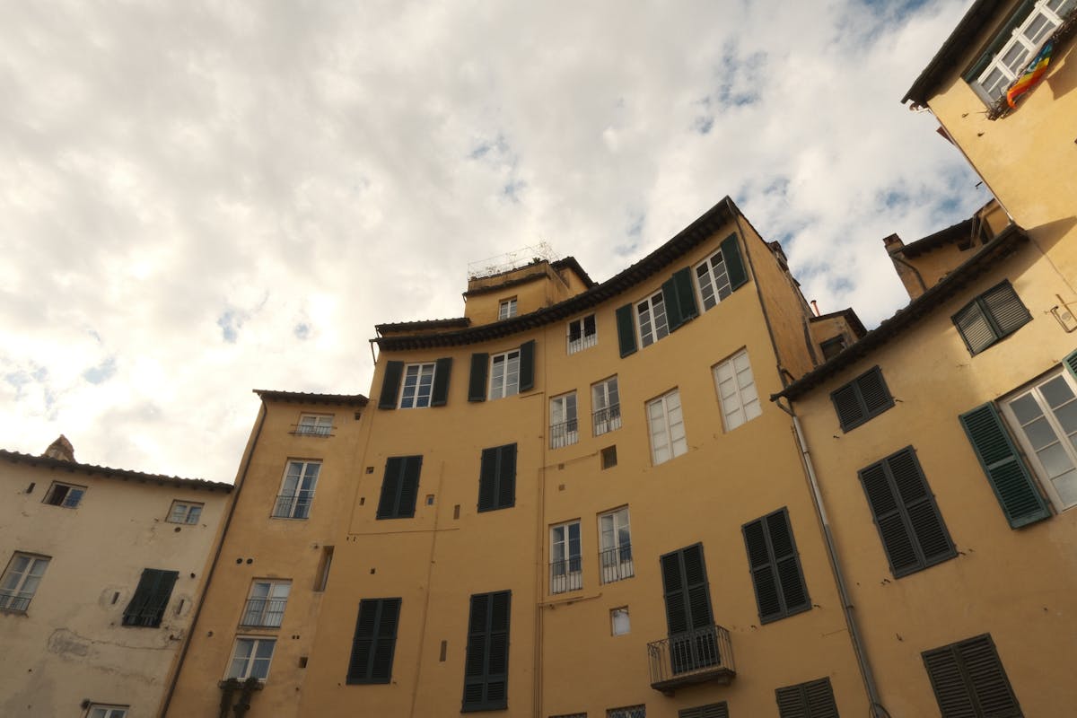 Curved yellow and ochre buildings following the elliptical line of Piazza dell'Anfiteatro in Lucca, with cloudy sky overhead