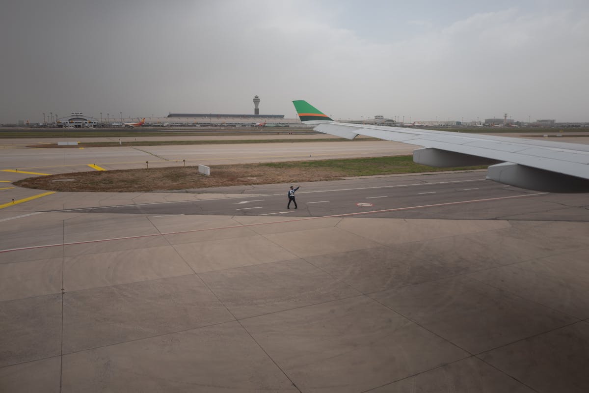 View of an airport tarmac with a ground marshaller directing a plane, terminal and control tower visible in the haze behind