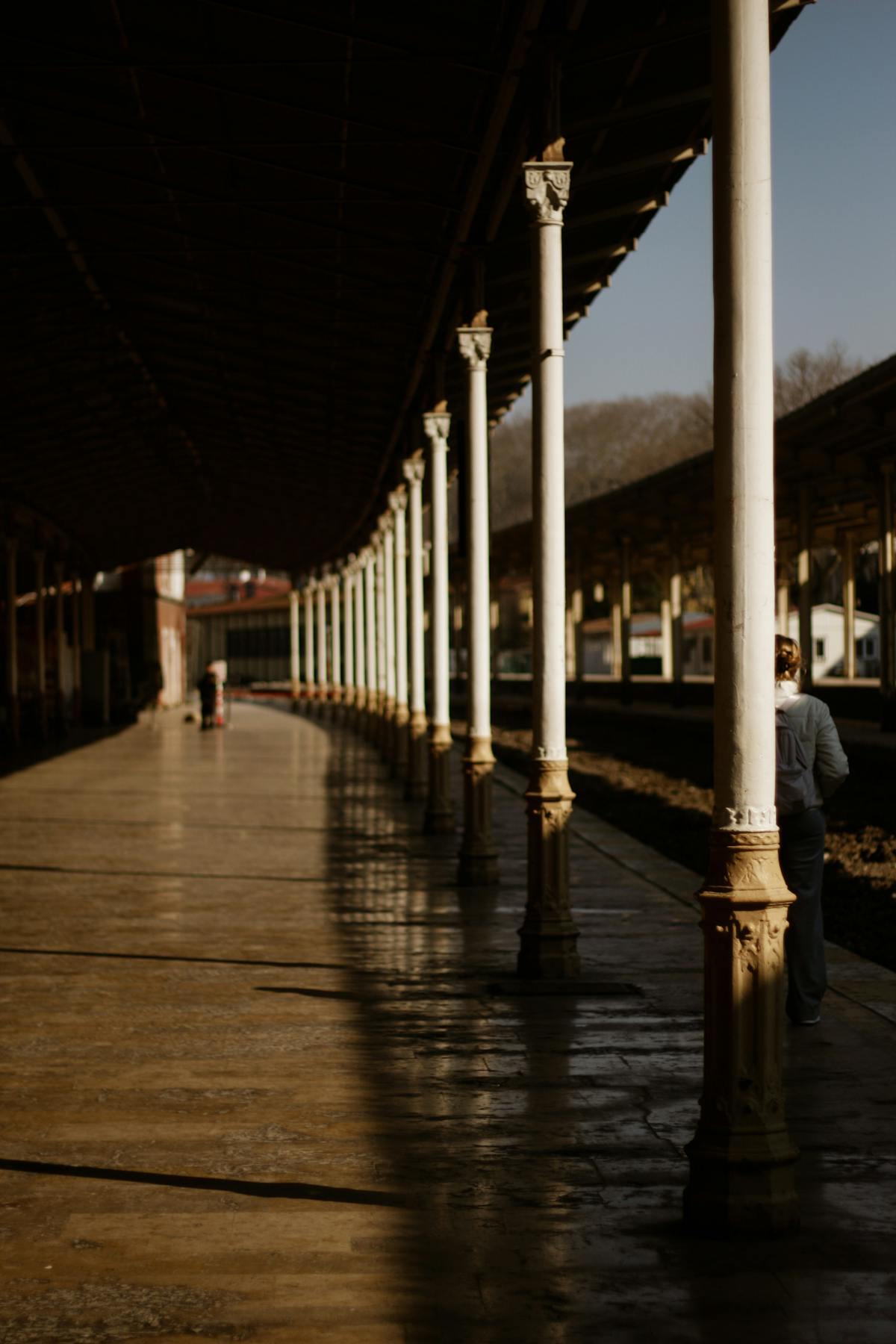 Long covered platform of an Italian railway station in late sun, with rows of cast-iron pillars receding into the distance